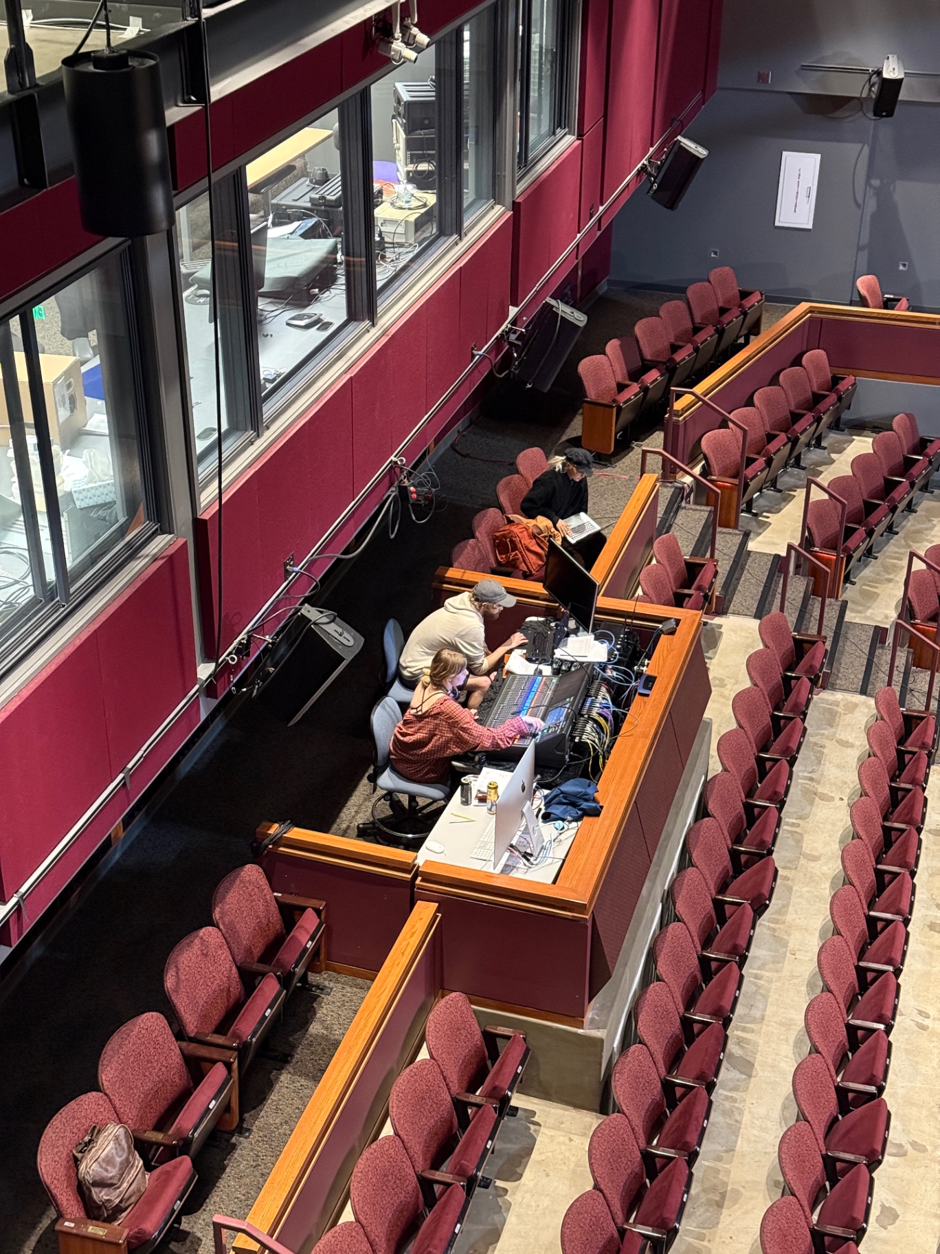 Overhead view of students working at a front-of-house mix position in a proscenium auditorium, with a digital console, monitors, and the control booth window behind them.