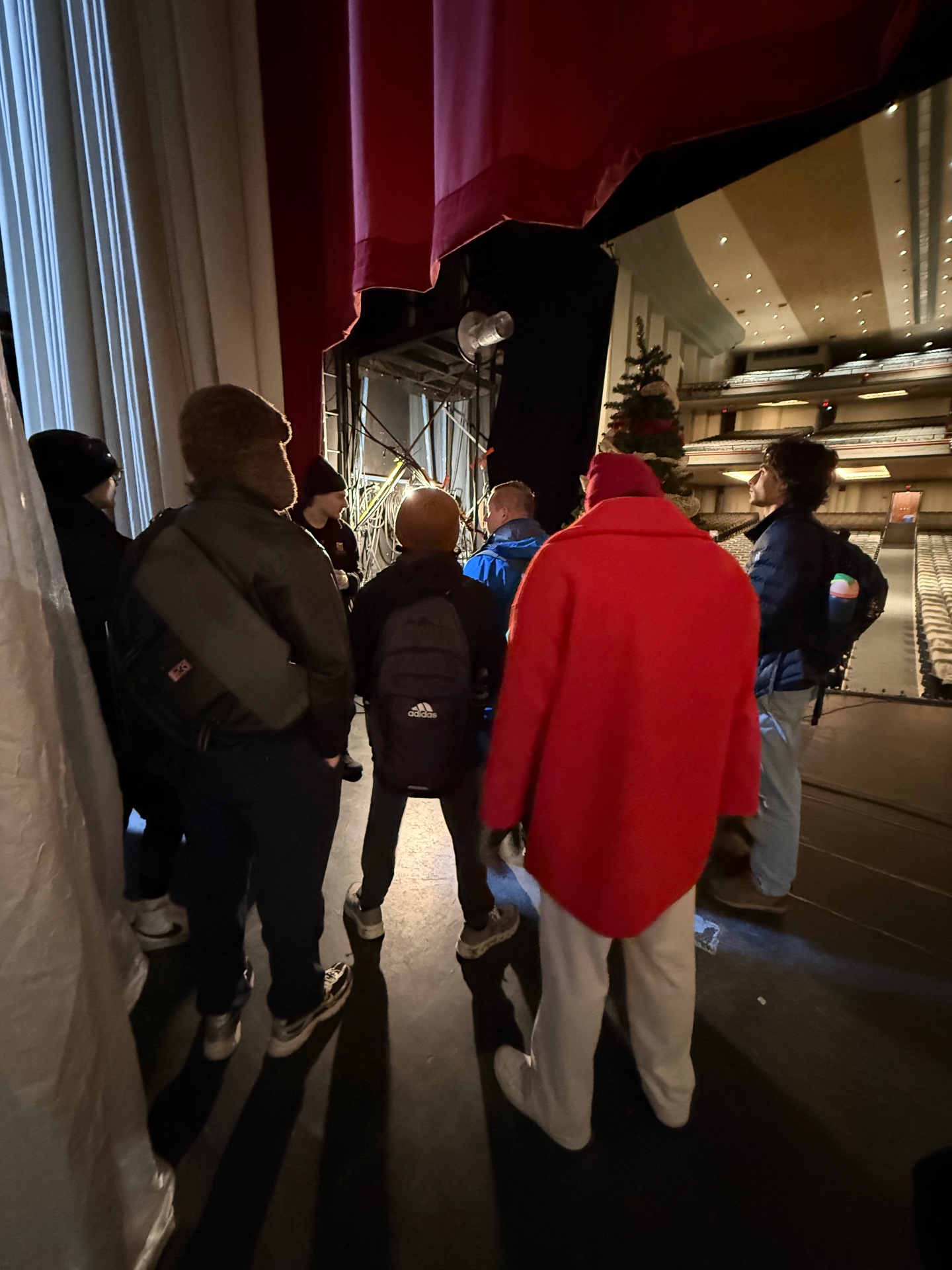 Students gathered upstage behind a red house curtain during a backstage tour of a large performing-arts venue, looking out at the empty house.