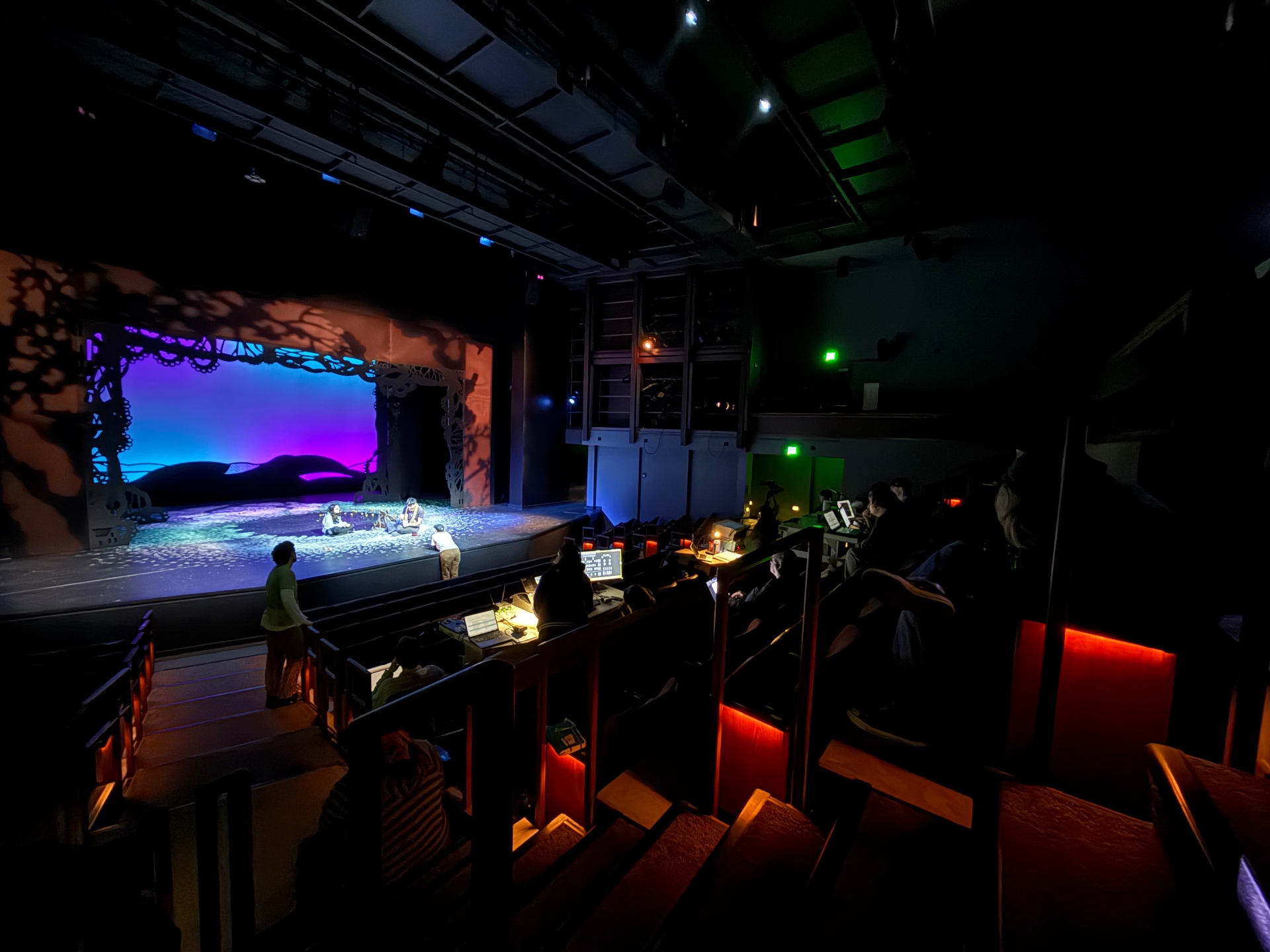 Tech rehearsal of a musical with a tree-silhouette set lit in deep purple and magenta, viewed from the back of the house.