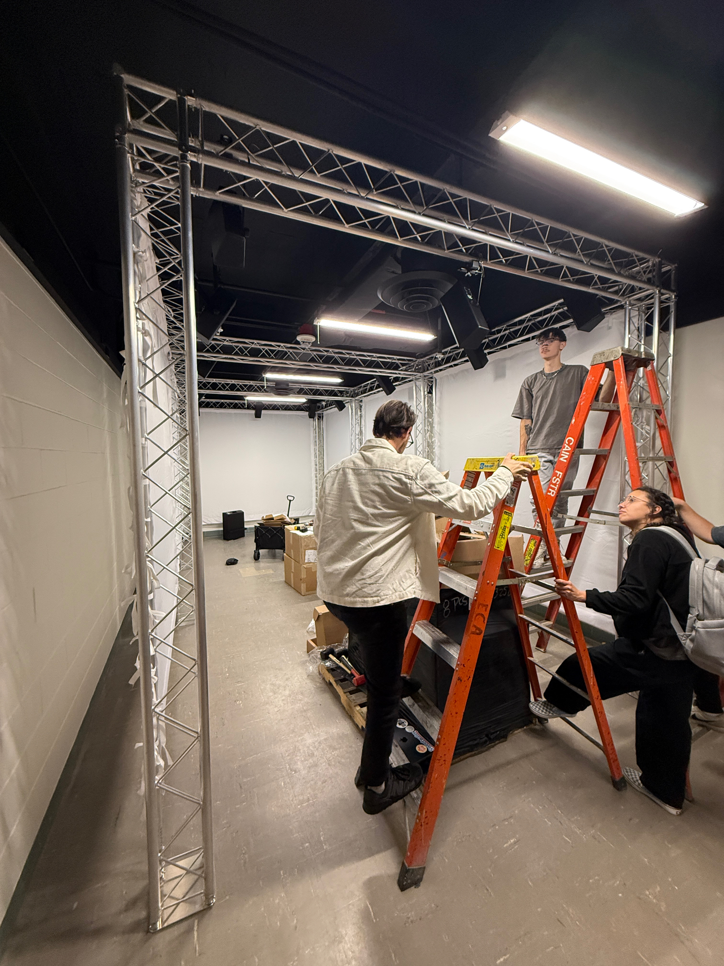 Students assembling and leveling a box-truss goalpost structure on ladders while building out a black-box studio space.