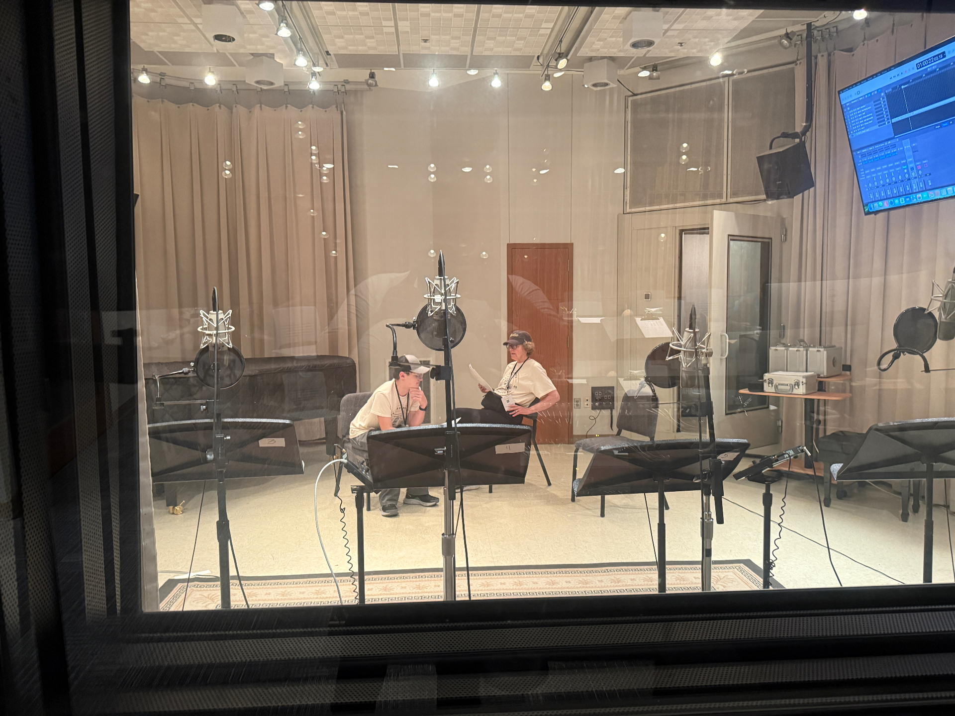 View through the control-room glass into the live room during a session, with two voice actors at music stands seated among condenser microphones in shock mounts.
