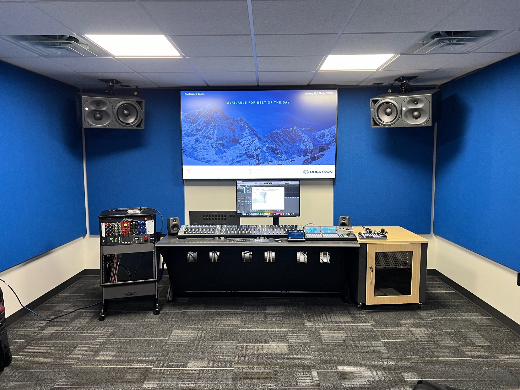 Completed audio control room with a digital mixing console, outboard rack, studio monitors, and a large display running room scheduling.